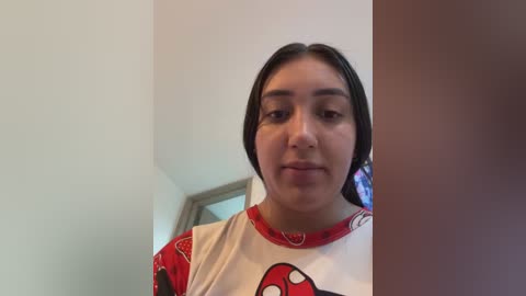 Media: Video of a young Latina woman with light skin, long dark hair, and a slight smile, wearing a red and white Spider-Man shirt. The background shows a plain, white wall with a door and window frame.