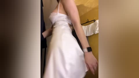 Video of a fair-skinned woman in a white satin slip dress, wearing a smartwatch, standing in a dimly lit room with beige walls and a white shirt hanging on a hook.