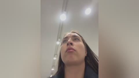 Media: Video of a woman with straight brown hair, wearing a black jacket, gazing upwards, under bright ceiling lights in a modern, minimalist interior.