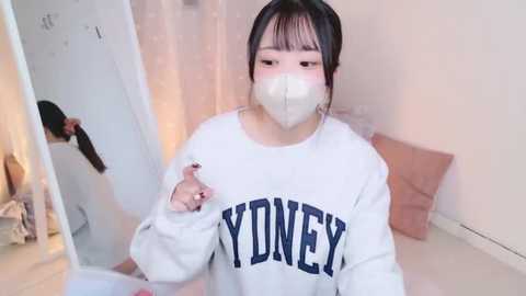 Media: Video of an Asian woman with dark hair and fair skin, wearing a white \"YONEY\" sweatshirt, face mask, and earrings, in a cozy, softly lit bedroom with pink pillows and a mirror.