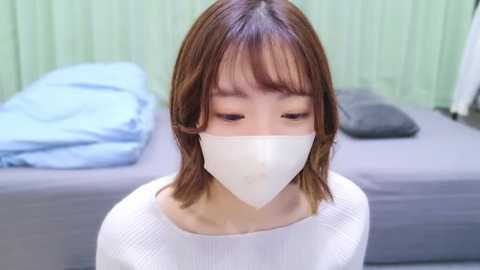 Media: Video of a young Asian woman with shoulder-length brown hair, wearing a white ribbed sweater and a white face mask, standing in a bedroom with a gray bed and blue blanket.