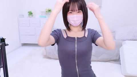 Media: A young Asian woman with straight black hair, wearing a gray shirt, face mask, and gloves, adjusts her hair in a minimalist, white room with a bed and white dresser.