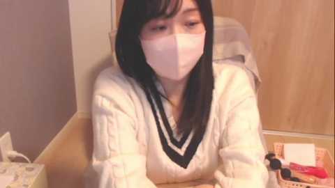 Media: A video of an Asian woman with long black hair, wearing a white sweater, a face mask, and a beige headband, seated in a wooden chair, in a room with light-colored walls and a desk with papers and a red book.