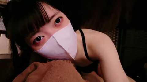 Media: Video of an Asian woman with long black hair, wearing a white surgical mask, black tank top, and a beige blanket, in a dimly lit room, appearing distressed.