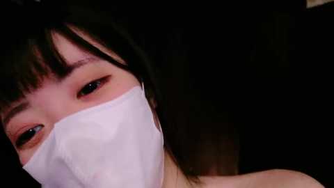 Media: Close-up video of an Asian woman with short black hair and pale skin, partially obscured by a white surgical mask, eyes visible, dark background.