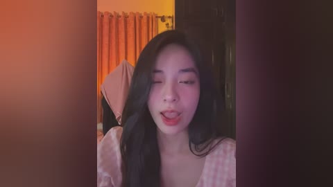 Media: Video of a young Asian woman with long black hair, wearing a white and pink polka-dotted shirt, smiling and sticking out her tongue in a playful manner. Background includes orange curtains and dark furniture.