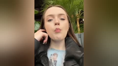 Media: A video of a young woman with fair skin and long brown hair, wearing a black jacket and a light blue shirt with a graphic, kissing her hand, set against lush greenery.