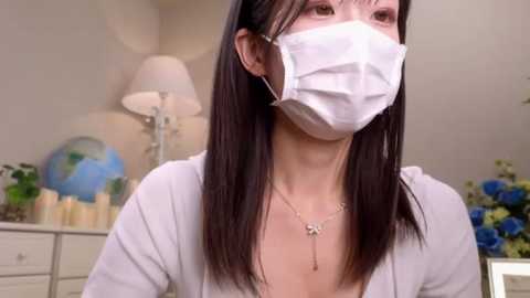 Media: Video of an Asian woman with long black hair, wearing a white mask, white top, and necklace, in a bright room with a globe, flowers, and candles on a white dresser.