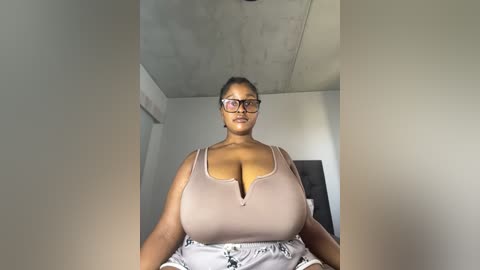 Media: A video of a plus-sized Black woman with a voluptuous figure, wearing a low-cut, beige tank top and patterned skirt, standing indoors with a minimalist, concrete ceiling and white walls.