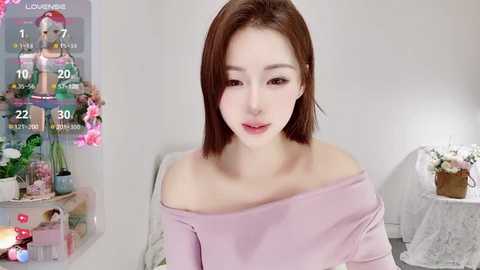 Media: Video of a young East Asian woman with fair skin and straight brown hair, wearing a light pink off-shoulder top, sitting indoors with a floral backdrop and a candlelit table.