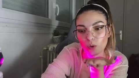 Media: Video of a young woman with glasses, light skin, and long hair in a ponytail, using a pink glowing vape pen indoors.