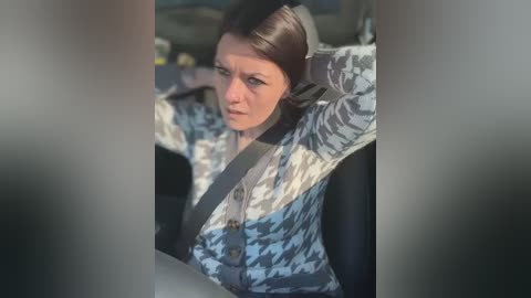 Media: A video shows a woman with brown hair, wearing a patterned jacket, adjusting her seat belt while driving. The background is blurred, with a hint of a dark car interior.