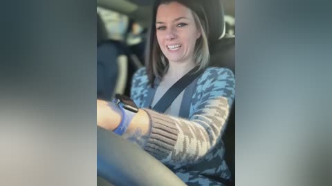 Media: Video of a smiling Caucasian woman with shoulder-length blonde hair, wearing a blue camo jacket and gray sweater, driving a car, with a black seatbelt and a blue wristband.