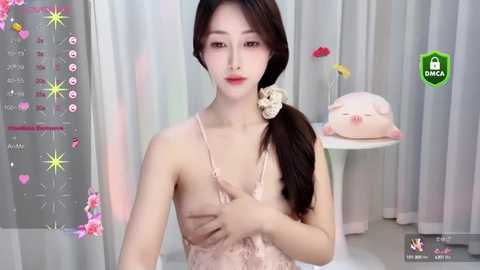 Media: Video of a slender Asian woman with long dark hair, wearing a pale pink lace dress, sitting against a white curtain background.