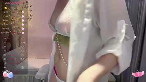 Media: A video shows a woman in a sheer white robe, revealing a green bra and pink underwear. The background features a blurred, cozy indoor setting with a wooden table and soft lighting.