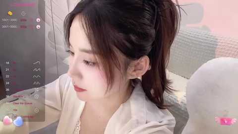 Media: Video of an Asian woman with fair skin and long dark hair in a high ponytail, wearing a white hoodie, sitting on a white couch.