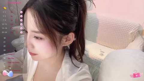 Media: Video of a young Asian woman with fair skin and long, dark hair in a ponytail, wearing a white shirt. She is sitting on a bed with a pastel-colored quilt and a stuffed animal. The background is soft pink and white.