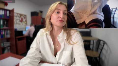 Media: A video of a blonde woman in a white button-up shirt, sitting at a desk in a cluttered office with bookshelves and a computer monitor.