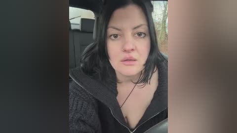 Media: Video of a woman with shoulder-length black hair, wearing a black hoodie, sitting in a car, looking serious. Background shows car seat and blurred outdoor scene.