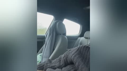 Media: Video of a passenger seat inside a car, with a grey, knitted blanket covering the passenger's lap. The passenger is wearing a dark hoodie and has long hair partially covering their face. The car's interior is dimly lit, and the background shows a blurred view of the road through the window.