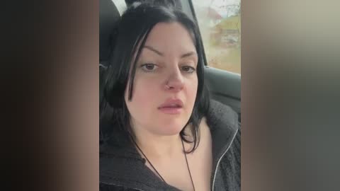 Media: Video of a woman with fair skin, black hair, and a black jacket, sitting in a car with a blurred, out-of-focus background.