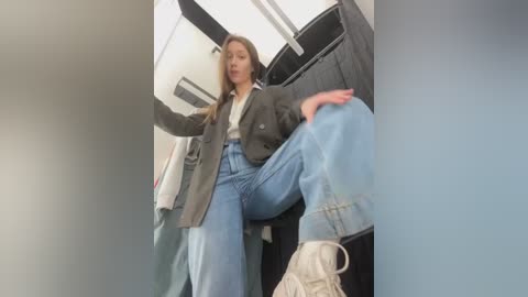 Media: Video of a young woman in a green blazer, light-wash jeans, and sneakers, sitting in a plane aisle, leaning forward, with a relaxed, casual demeanor.