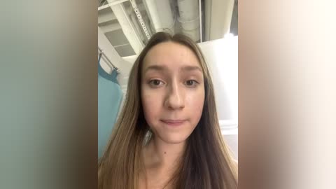 Media: Video of a young Caucasian woman with straight, light brown hair and fair skin, standing indoors. She wears a light-colored top. Background features white pipes and a blue wall.