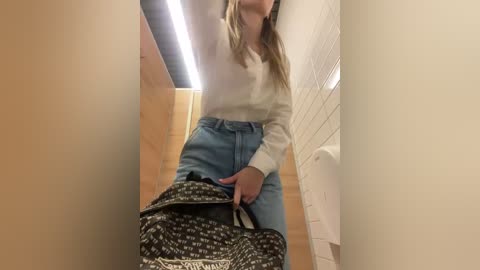 Media: A video of a woman in a bathroom stall, wearing a white sweater and high-waisted light blue jeans, carrying a black and white patterned handbag. The background features tiled walls and a flushable toilet.