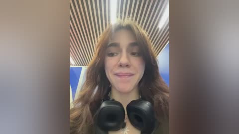 Media: Video of a young woman with long brown hair, wearing headphones, smiling. Background features a modern, blue and brown ceiling with vertical stripes.