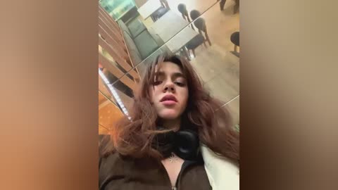 Media: A video of a young woman with wavy brown hair, wearing a black jacket and black headphones, sitting in a modern, glass-walled cafe with light wood furniture and green accents.