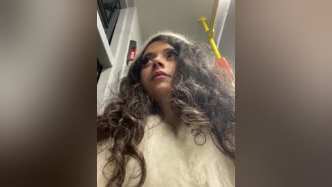 A video of a young woman with long, curly, dark hair, fair skin, and light makeup, standing in a subway car, looking up. She wears a cream-colored top. Background shows other passengers, subway signs, and yellow poles.