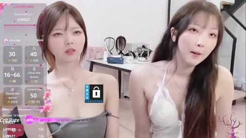 Media: Video of two young East Asian women with light skin, one in a gray strapless top and the other in a white lace bra, sitting indoors.