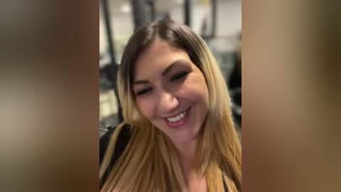 Media: Video of a smiling Asian woman with straight, long, blonde hair, wearing a black top, sitting in a blurred gym setting.