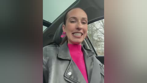 Media: Video of a smiling woman with light skin and short brown hair, wearing a bright pink turtleneck and a grey leather jacket, seated in a car with a blurred outdoor view.