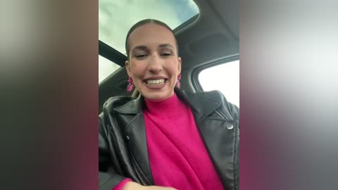 Media: Video of a smiling woman with straight, dark hair, wearing a bright pink turtleneck under a black leather jacket, sitting in a car.