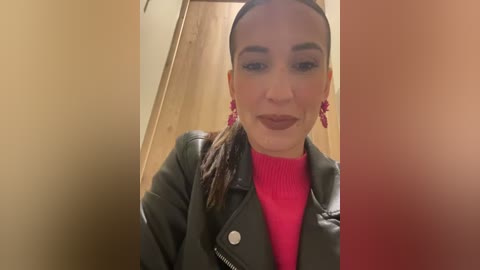 Media: Video of a young woman with fair skin and dark hair styled in a high ponytail. She wears a green leather jacket over a bright pink sweater, red earrings, and dark lipstick. The background is a beige wall and wooden door.