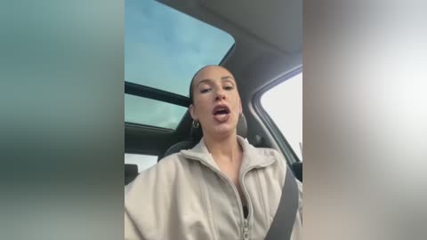 Media: Video of a light-skinned woman with short hair, wearing a beige jacket, driving a car with a sunroof visible. She has her mouth open, possibly in mid-speech.