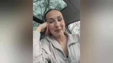 Media: Video of a young woman with light skin, brown eyes, and dark hair pulled back, wearing a light gray hoodie, sitting in a car with a sunroof, looking relaxed.