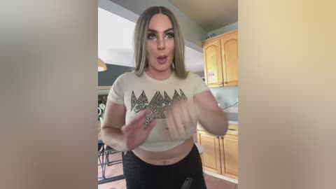 Media: A video of a curvy, light-skinned woman with shoulder-length blonde hair, wearing a cropped white T-shirt with geometric patterns, and black pants, standing in a kitchen with light wood cabinets.