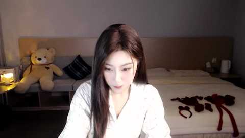 Media: A video of a young Asian woman with long black hair, fair skin, and medium-sized breasts, wearing a white robe, sitting in a dimly-lit bedroom with a teddy bear, candles, and a neatly made bed.