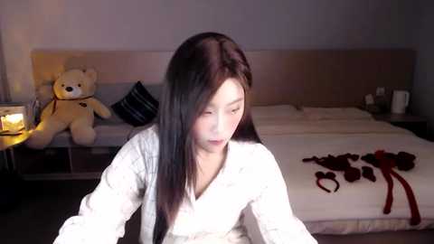 Media: A video of a young Asian woman with long black hair, wearing a white robe, sitting on a bed with teddy bears, candles, and red lingerie scattered on the white sheets.