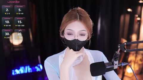 Media: Video of an Asian woman with fair skin, brown hair, and glasses, wearing a black face mask and white blouse, singing into a microphone. Background shows dimly lit stage with blurred audience.