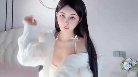 Media: Video of an Asian woman with long, straight black hair, fair skin, and large breasts, wearing a white robe, posing in a modern bedroom with white walls and a white tufted headboard.