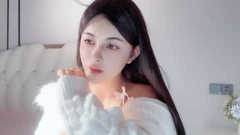 Media: Video of a young Asian woman with long black hair, wearing a white off-shoulder sweater, standing in a modern, minimalist room with a white couch and a wall-mounted light.