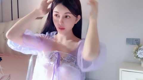 Media: Video of an East Asian woman with long black hair, wearing a sheer, lavender, ruffled top, adjusting a headband in a softly lit, minimalist bedroom with white walls.