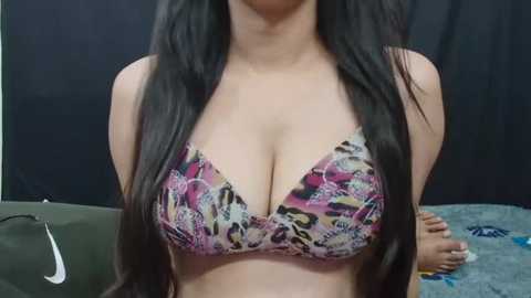 Media: Video of a woman with long, straight black hair, wearing a colorful leopard print bikini top that accentuates her ample cleavage. She stands against a dark background with a green couch visible on the left.