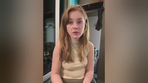 Video of a young Caucasian woman with fair skin, long blonde hair, and a neutral expression, wearing a beige turtleneck sweater and high-waisted pants. Background features a cluttered room with hanging clothes and a partially visible window.