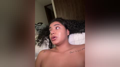 Media: Video of a topless, light-skinned woman with curly hair, tattoos on her shoulders, looking surprised and slightly open-mouthed, in a bathroom with white tiled walls and wooden cabinets.