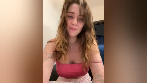 Media: Video of a young woman with long, wavy brown hair and light skin, wearing a pink tank top and a pink skirt. She has tattoos on her arms and is smiling softly.