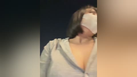 Media: A video of a woman with long dark hair and a white surgical mask covering her mouth, wearing a low-cut, light-colored shirt, set against a dark background.